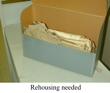 Unorganized newspaper clippings in archival box with caption "Rehousing needed".